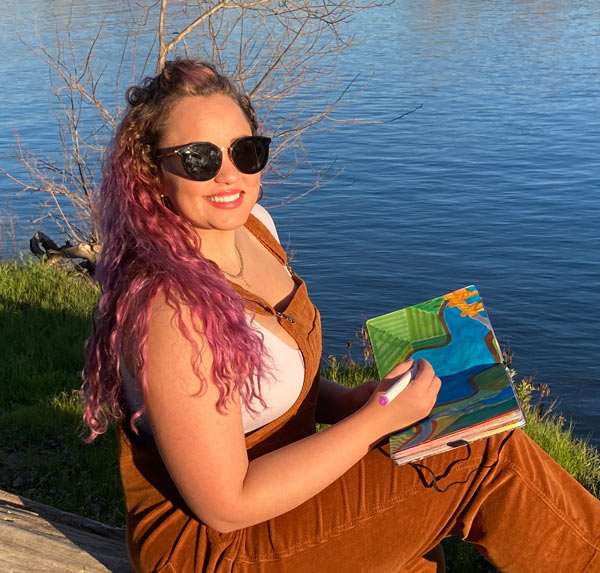 Rachael Prato sitting by the rivers edge with sunglasses on sketching the mural in her notebook with a marker