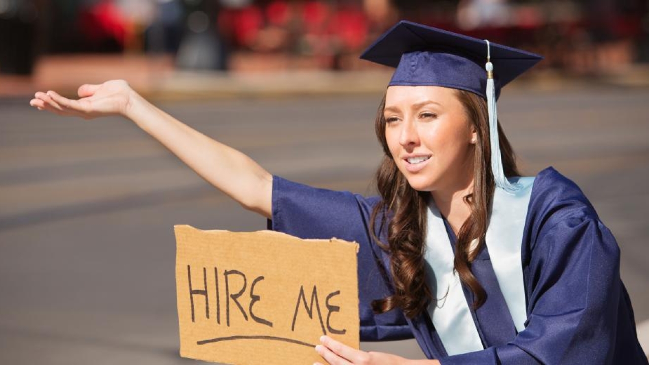 Woman in graduation gown and cap holding out a cardboard sign that has "hire me" written in black marker as she tries to wave down cars passing by.