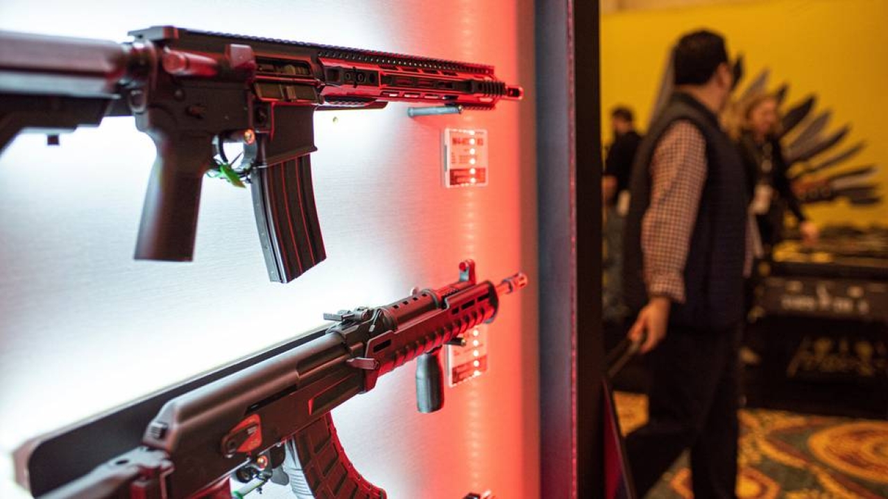 In the foreground, there are two guns hanging on a wall, the top appearing to be a semi automatic and the bottom an automatic rifle. To the right of this wall in the background are some people blurred out potentially at a convention.