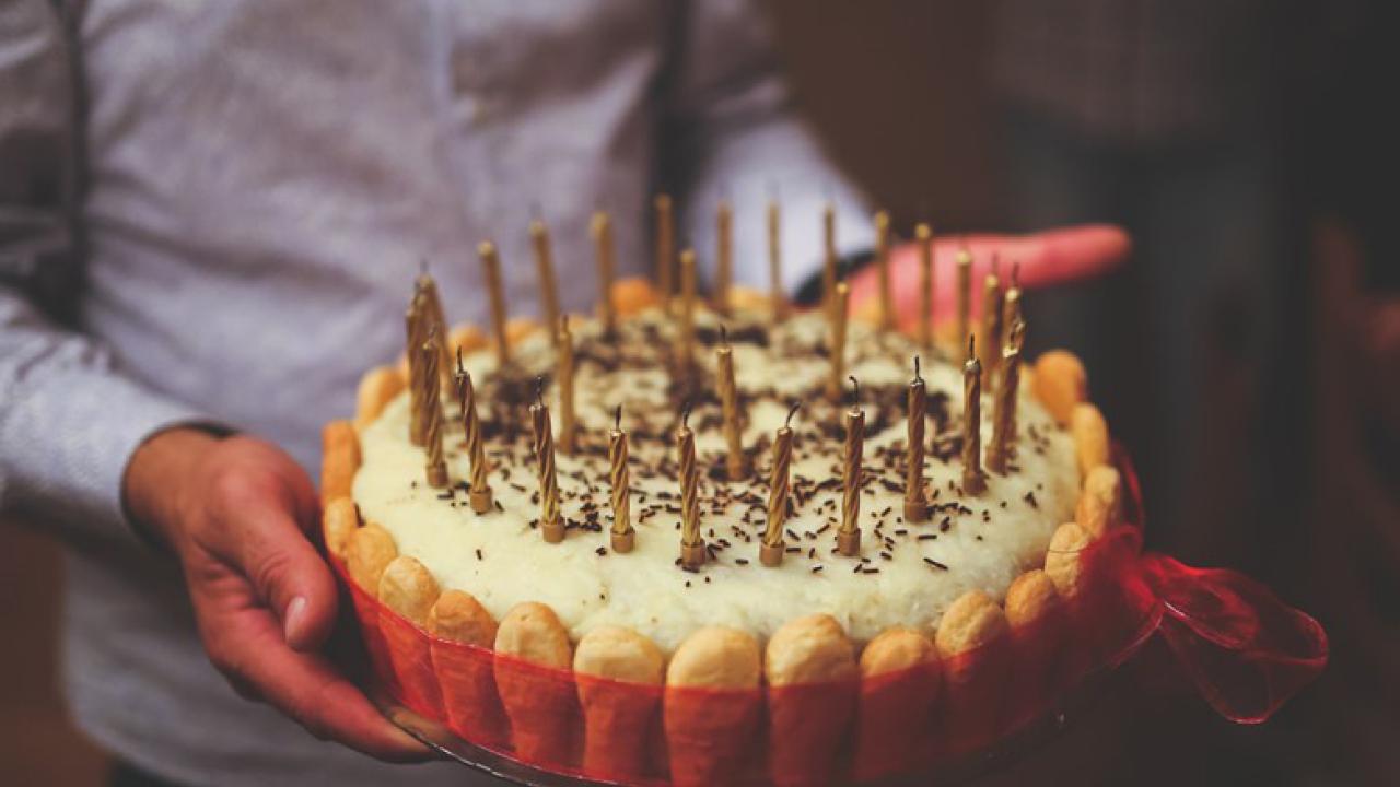 Person holding a tiramisu cake with birthday candles on top. Cake is wrapped in red ribbon.