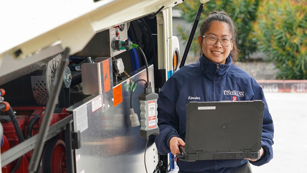 Kimmy Nguyen standing next to bus with open compartment exposing mechanical workings while holding a computer and wearing her unitrans jacket.