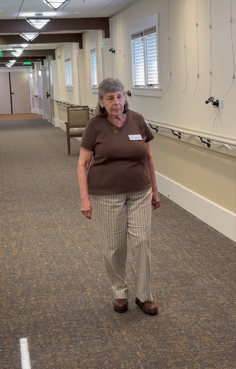A retirement home resident walking down the hallway wearing a brown shirt, nametag and striped pants.