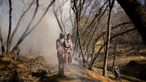 Two women on a hillside with smoke behind them during prescribed burn.