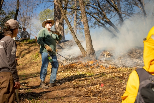 Man with beard and hat talks to group in wooded clearing during a prescribed burn.