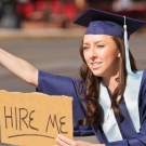 Woman in graduation gown and cap holding out a cardboard sign that has "hire me" written in black marker as she tries to wave down cars passing by.