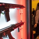 In the foreground, there are two guns hanging on a wall, the top appearing to be a semi automatic and the bottom an automatic rifle. To the right of this wall in the background are some people blurred out potentially at a convention.