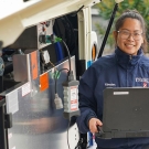 Kimmy Nguyen standing next to bus with open compartment exposing mechanical workings while holding a computer and wearing her unitrans jacket.